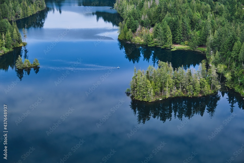 Person on paddle board on the calm lake with islands and reflections in water. View from above. Lily lake in Madeira Park. Sunshine Coast. British Columbia. Canada Person on paddle board on the calm lake with islands and reflections in water. View from above. Lily lake in Madeira Park. Sunshine Coast. British Columbia. Canada