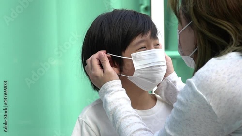 Close-up of an Asian mother putting a protective medical face mask on her little son outdoors.