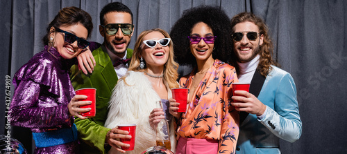 smiling interracial friends in sunglasses standing on grey background, banner.