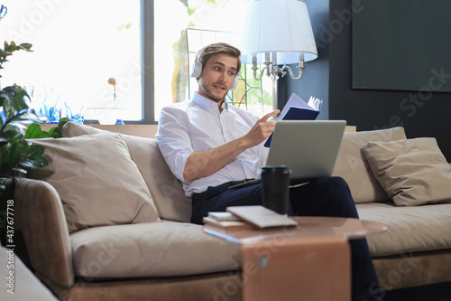 Concentrated young freelancer businessman sitting on sofa with laptop and examining documents.