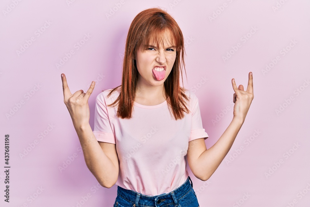 Redhead young woman wearing casual pink t shirt shouting with crazy expression doing rock symbol with hands up. music star. heavy concept. Redhead young woman wearing casual pink t shirt shouting with crazy expression doing rock symbol with hands up. music star. heavy concept.