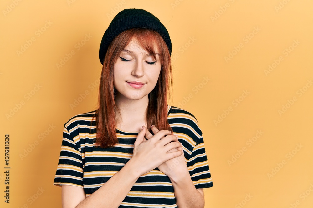 Redhead young woman wearing wool cap smiling with hands on chest with closed eyes and grateful gesture on face. health concept.