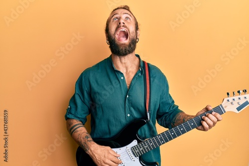 Handsome man with beard and long hair playing electric guitar angry and mad screaming frustrated and furious, shouting with anger looking up.