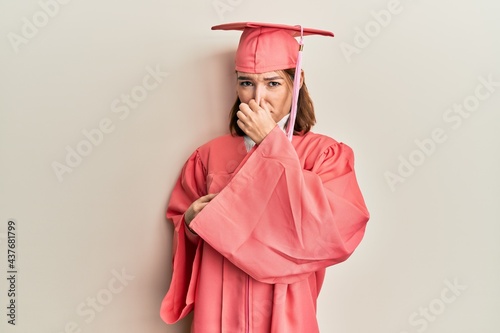 Young caucasian woman wearing graduation cap and ceremony robe smelling something stinky and disgusting, intolerable smell, holding breath with fingers on nose. bad smell