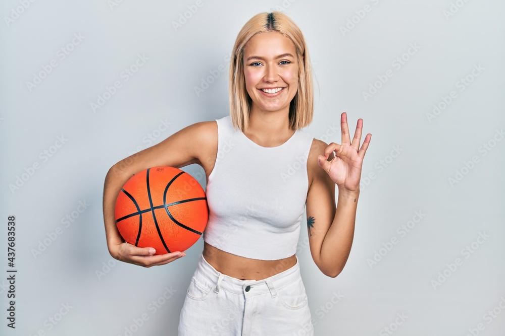 Beautiful blonde woman holding basketball ball doing ok sign with fingers, smiling friendly gesturing excellent symbol Beautiful blonde woman holding basketball ball doing ok sign with fingers, smiling friendly gesturing excellent symbol