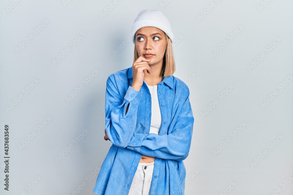Beautiful blonde woman wearing wool hat with hand on chin thinking about question, pensive expression. smiling with thoughtful face. doubt concept. Beautiful blonde woman wearing wool hat with hand on chin thinking about question, pensive expression. smiling with thoughtful face. doubt concept.