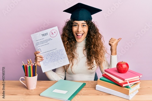 Young hispanic girl wearing graduated hat holding passed test pointing thumb up to the side smiling happy with open mouth photo