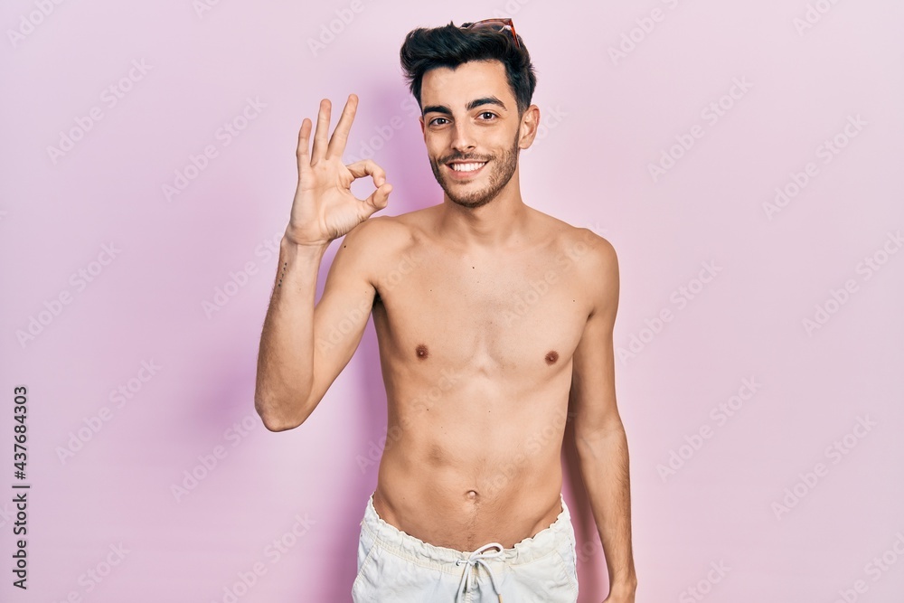 Young hispanic man wearing swimwear shirtless smiling positive doing ok sign with hand and fingers. successful expression.