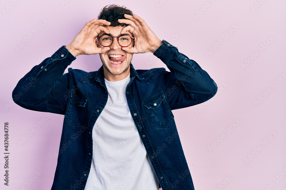 Young hispanic man wearing casual clothes and glasses doing ok gesture like binoculars sticking tongue out, eyes looking through fingers. crazy expression. Young hispanic man wearing casual clothes and glasses doing ok gesture like binoculars sticking tongue out, eyes looking through fingers. crazy expression.