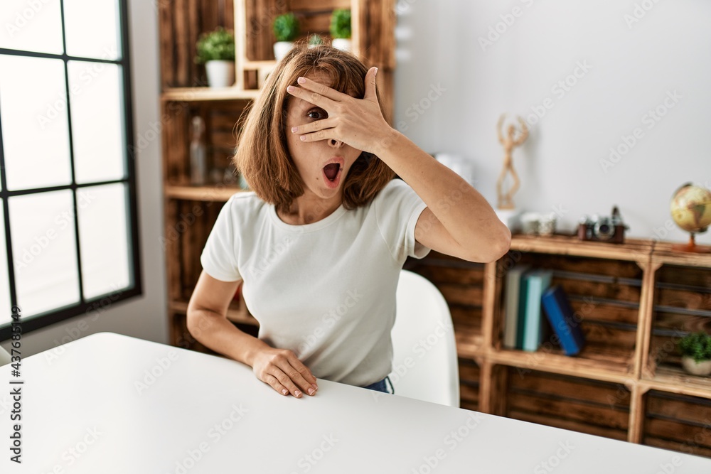 Young caucasian girl wearing casual clothes sitting on the table at home peeking in shock covering face and eyes with hand, looking through fingers with embarrassed expression. Young caucasian girl wearing casual clothes sitting on the table at home peeking in shock covering face and eyes with hand, looking through fingers with embarrassed expression.