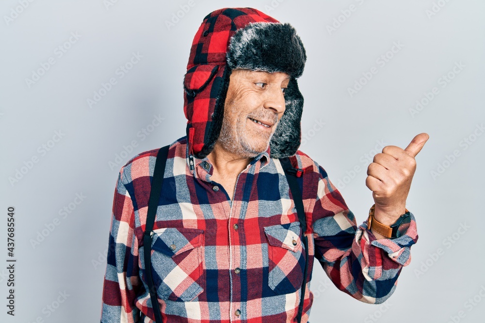 Naklejka premium Handsome mature man wearing winter hat with ear flaps smiling with happy face looking and pointing to the side with thumb up.
