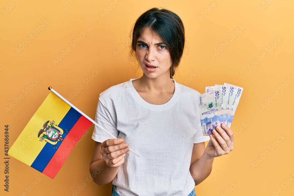 Young caucasian woman holding colombia flag and colombian pesos banknotes in shock face, looking skeptical and sarcastic, surprised with open mouth Young caucasian woman holding colombia flag and colombian pesos banknotes in shock face, looking skeptical and sarcastic, surprised with open mouth
