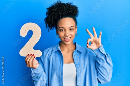 Young african american girl holding number two doing ok sign with fingers, smiling friendly gesturing excellent symbol