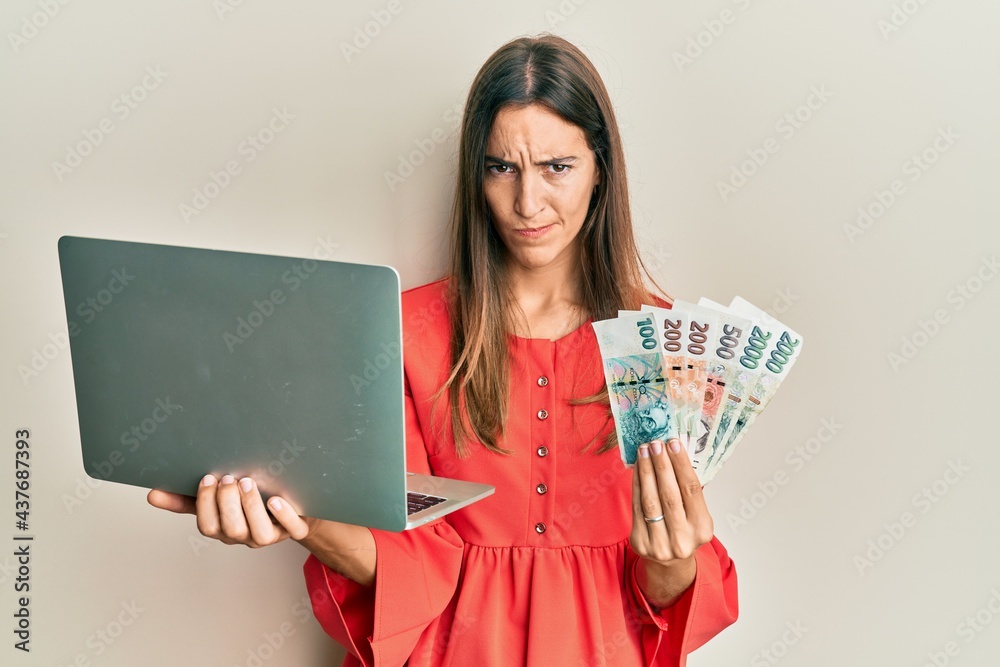 Young beautiful woman holding laptop and czech koruna banknotes skeptic and nervous, frowning upset because of problem. negative person. Young beautiful woman holding laptop and czech koruna banknotes skeptic and nervous, frowning upset because of problem. negative person.