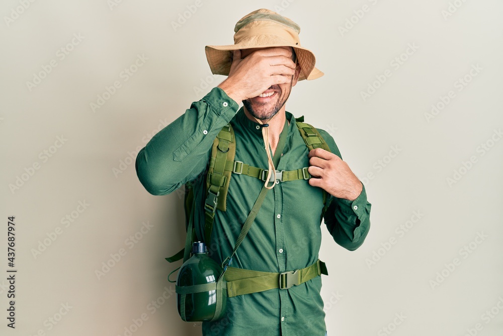 Handsome man with beard wearing explorer hat and backpack smiling and laughing with hand on face covering eyes for surprise. blind concept. Handsome man with beard wearing explorer hat and backpack smiling and laughing with hand on face covering eyes for surprise. blind concept.