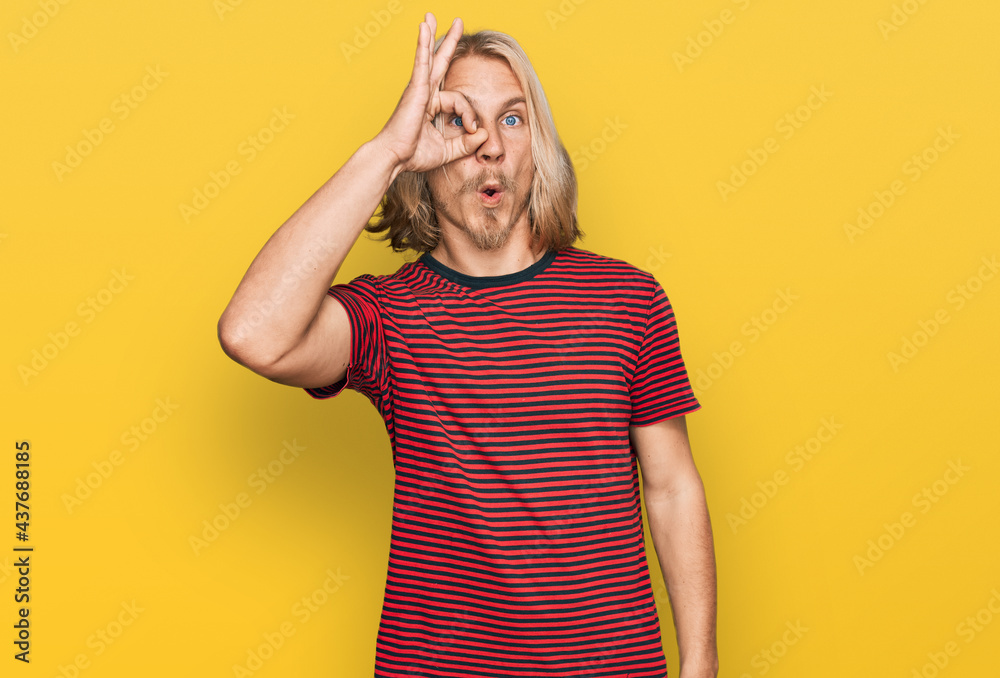 Caucasian man with blond long hair wearing casual striped t shirt doing ok gesture shocked with surprised face, eye looking through fingers. unbelieving expression. Caucasian man with blond long hair wearing casual striped t shirt doing ok gesture shocked with surprised face, eye looking through fingers. unbelieving expression.