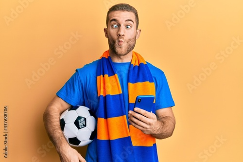 Young caucasian man holding football ball looking at smartphone making fish face with mouth and squinting eyes  crazy and comical.