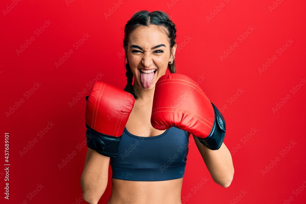 Young brunette girl using boxing gloves sticking tongue out happy with funny expression.