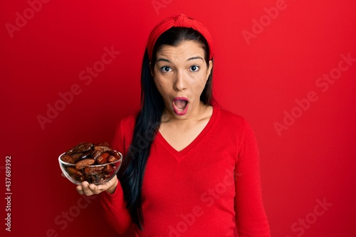 Young latin woman holding bowl with dates scared and amazed with open mouth for surprise  disbelief face
