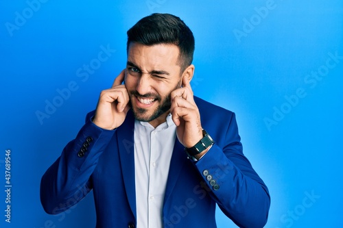 Young hispanic businessman wearing business jacket covering ears with fingers with annoyed expression for the noise of loud music. deaf concept.