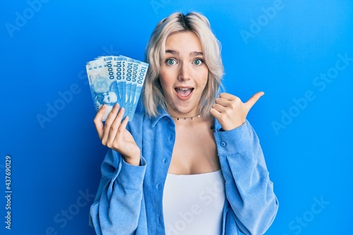 Young blonde girl holding 1000 chilean pesos pointing thumb up to the side smiling happy with open mouth