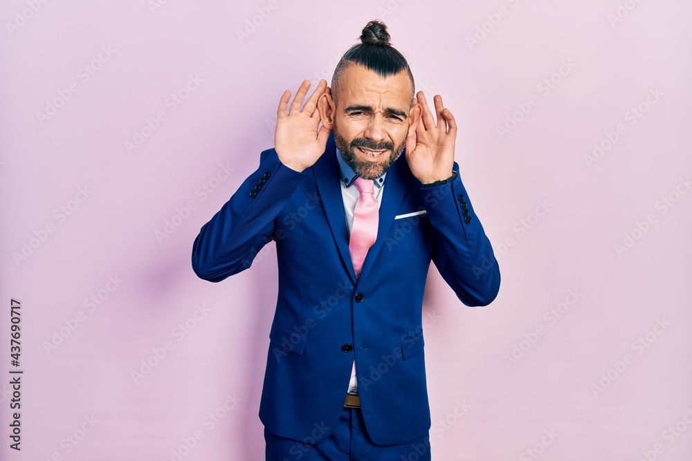 Young hispanic man wearing business suit and tie trying to hear both hands on ear gesture, curious for gossip. hearing problem, deaf Young hispanic man wearing business suit and tie trying to hear both hands on ear gesture, curious for gossip. hearing problem, deaf