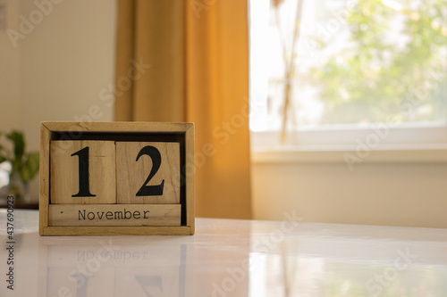 Wooden blocks of the calendar represents the date 12 and the month of November on the background of a window, curtain and a plant.