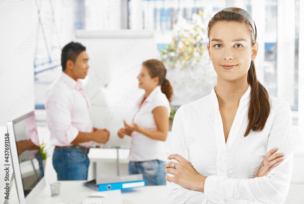 Fototapeta premium Portrait of young female office worker, looking at camera, smiling. 2 office workers in background talking.