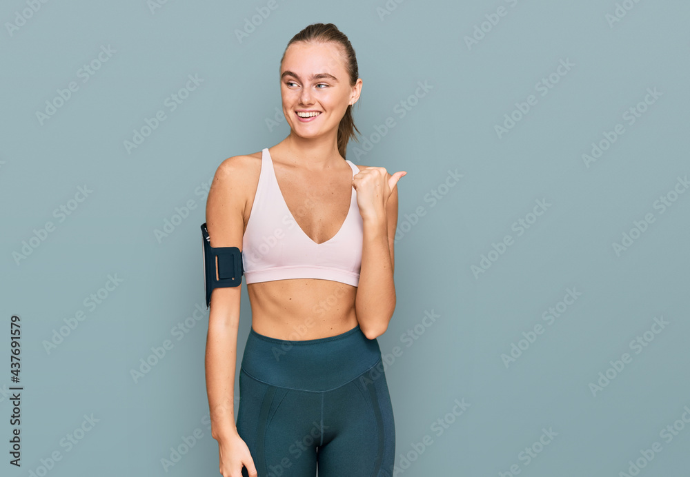 Beautiful young blonde woman wearing sportswear and arm band smiling with happy face looking and pointing to the side with thumb up. Beautiful young blonde woman wearing sportswear and arm band smiling with happy face looking and pointing to the side with thumb up.