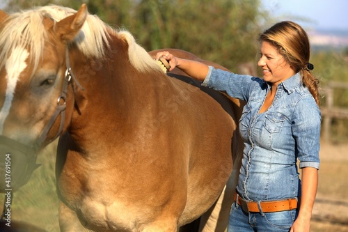 Happy young cowgirl standing next to horse, brushing, smiling.
