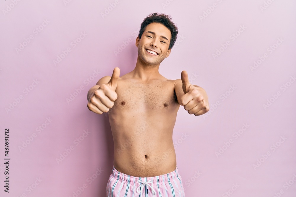 Young handsome man wearing swimwear shirtless approving doing positive gesture with hand, thumbs up smiling and happy for success. winner gesture.