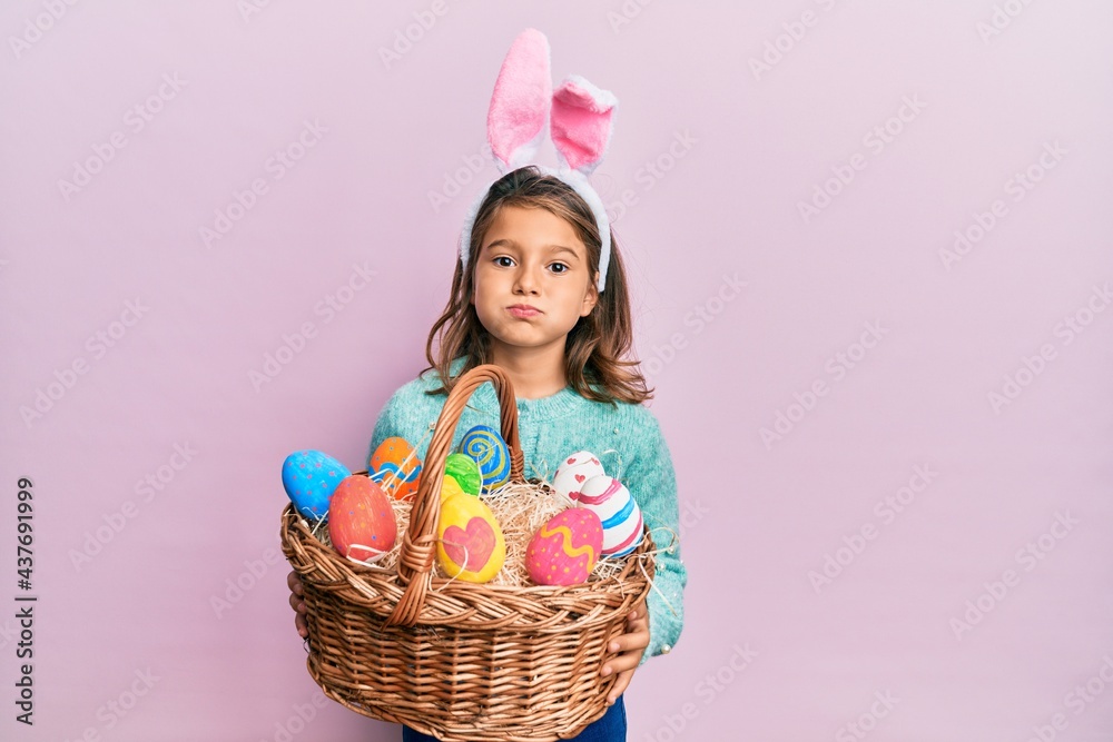 Little beautiful girl wearing cute easter bunny ears holding wicker basket with colored eggs puffing cheeks with funny face. mouth inflated with air, catching air.