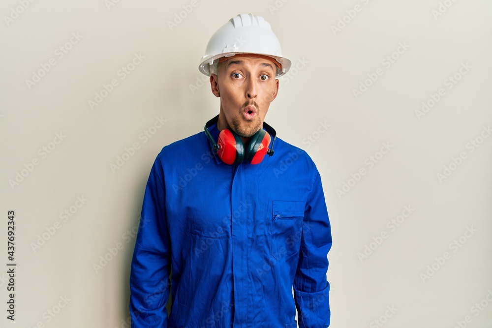 Bald man with beard wearing builder jumpsuit uniform and hardhat scared and amazed with open mouth for surprise, disbelief face