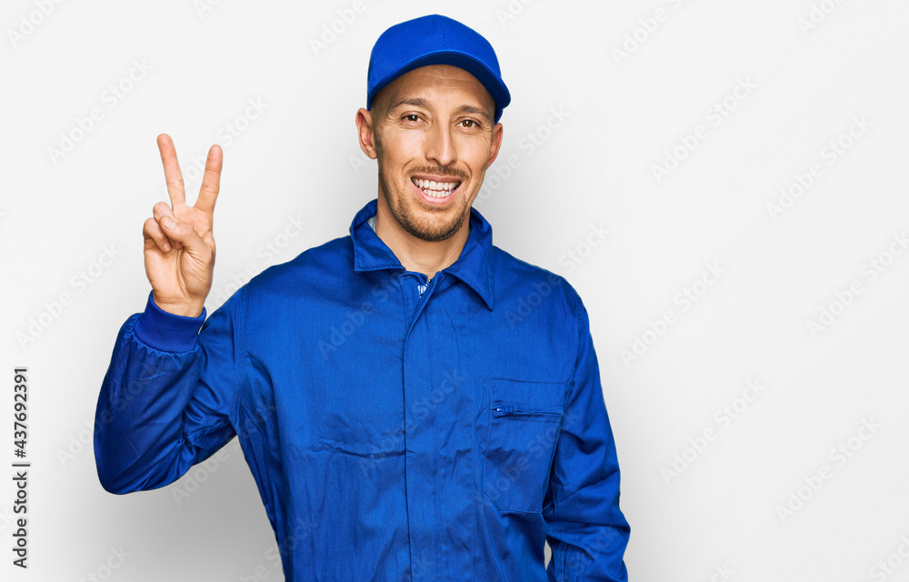 Bald man with beard wearing builder jumpsuit uniform smiling looking to the camera showing fingers doing victory sign. number two.