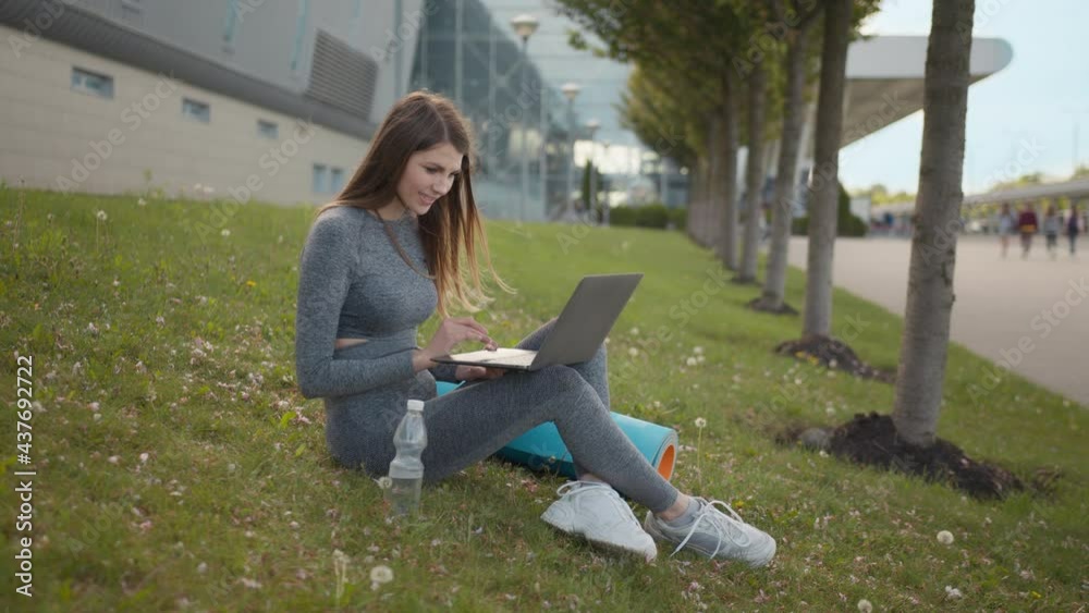 Portrait of fitness woman using laptop sitting on mat outdoors. Business lady working n laptop waiting for yoga class in summer park. Healthy lifestyle concept. Work And Training Portrait of fitness woman using laptop sitting on mat outdoors. Business lady working n laptop waiting for yoga class in summer park. Healthy lifestyle concept. Work And Training
