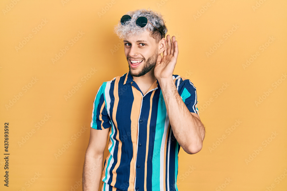 Young hispanic man with modern dyed hair wearing summer shirt and sunglasses smiling with hand over ear listening and hearing to rumor or gossip. deafness concept. Young hispanic man with modern dyed hair wearing summer shirt and sunglasses smiling with hand over ear listening and hearing to rumor or gossip. deafness concept.