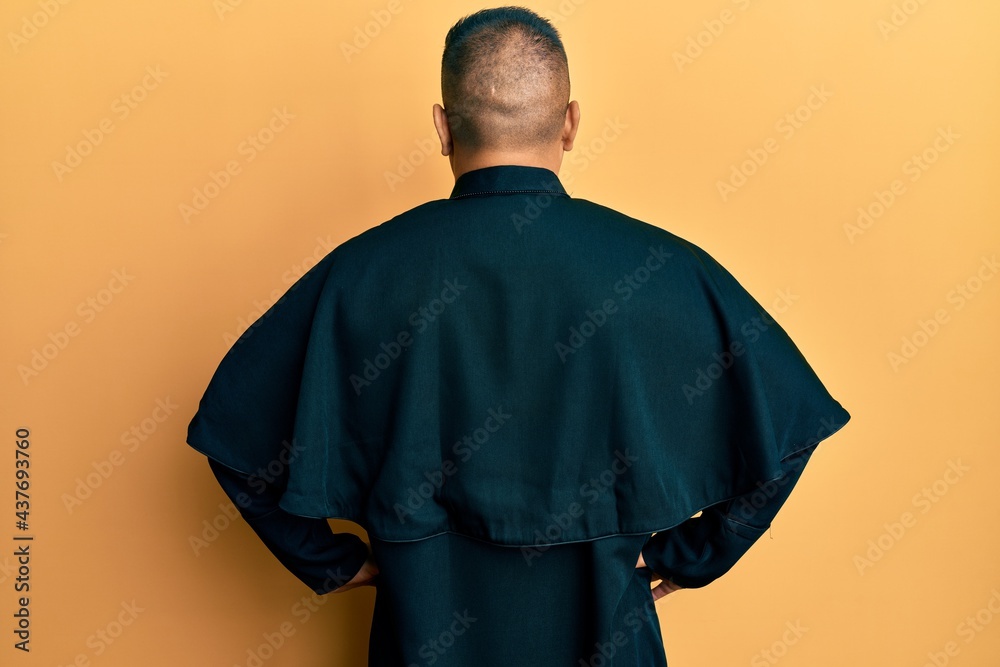 Obraz premium Young latin priest man standing over yellow background standing backwards looking away with arms on body