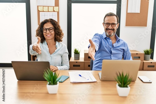 Middle age hispanic woman and man sitting with laptop at the office smiling friendly offering handshake as greeting and welcoming. successful business.