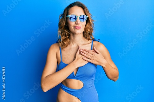 Young caucasian woman wearing swimsuit and sunglasses smiling with hands on chest, eyes closed with grateful gesture on face. health concept.