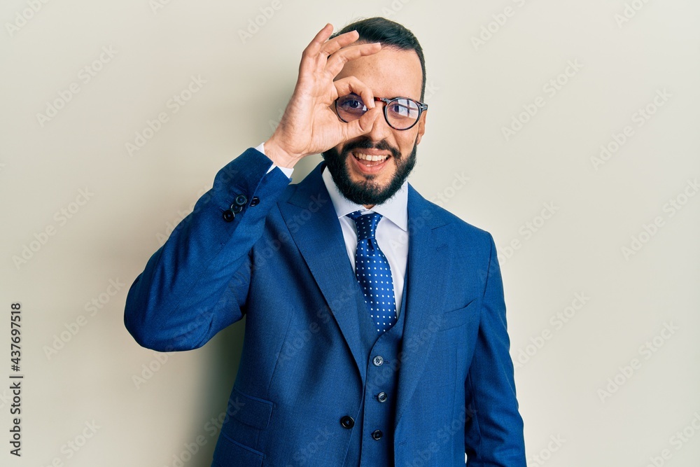 Young man with beard wearing business suit and tie doing ok gesture with hand smiling, eye looking through fingers with happy face. Young man with beard wearing business suit and tie doing ok gesture with hand smiling, eye looking through fingers with happy face.