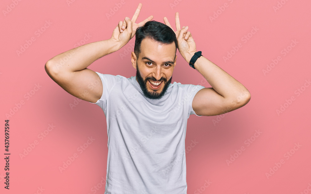 Young man with beard wearing casual white t shirt posing funny and crazy with fingers on head as bunny ears, smiling cheerful Young man with beard wearing casual white t shirt posing funny and crazy with fingers on head as bunny ears, smiling cheerful
