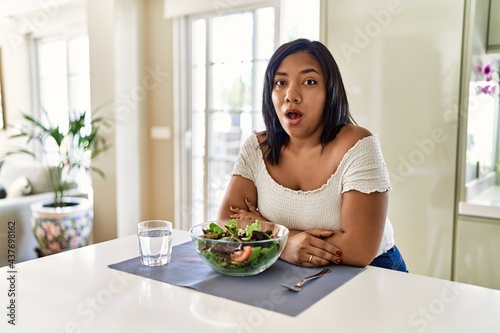 Young hispanic woman eating healthy salad at home afraid and shocked with surprise expression, fear and excited face.