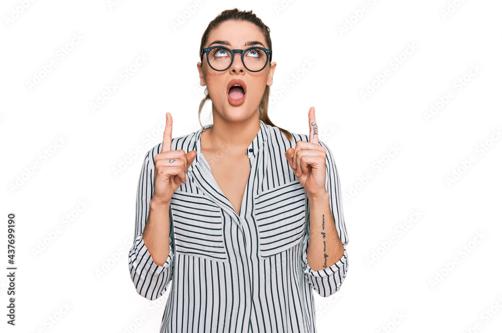 Young caucasian woman wearing business shirt and glasses amazed and surprised looking up and pointing with fingers and raised arms. Young caucasian woman wearing business shirt and glasses amazed and surprised looking up and pointing with fingers and raised arms.