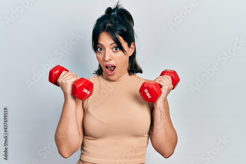Young hispanic woman wearing sportswear using dumbbells in shock face, looking skeptical and sarcastic, surprised with open mouth