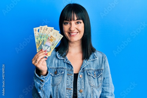 Young hispanic woman holding 50 romanian leu banknotes looking positive and happy standing and smiling with a confident smile showing teeth