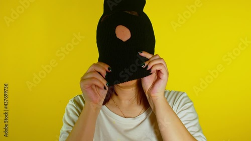 Close up young woman puts on black balaclava on yellow background. Secretive female puts on mask, looking at camera. photo