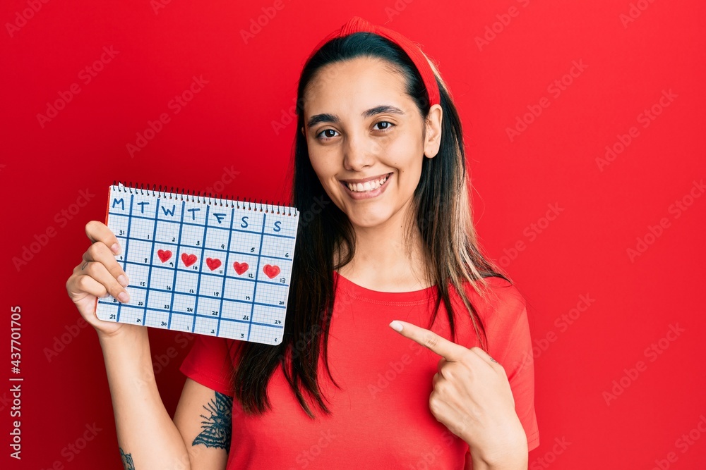 Young hispanic woman holding heart calendar smiling happy pointing with hand and finger Young hispanic woman holding heart calendar smiling happy pointing with hand and finger
