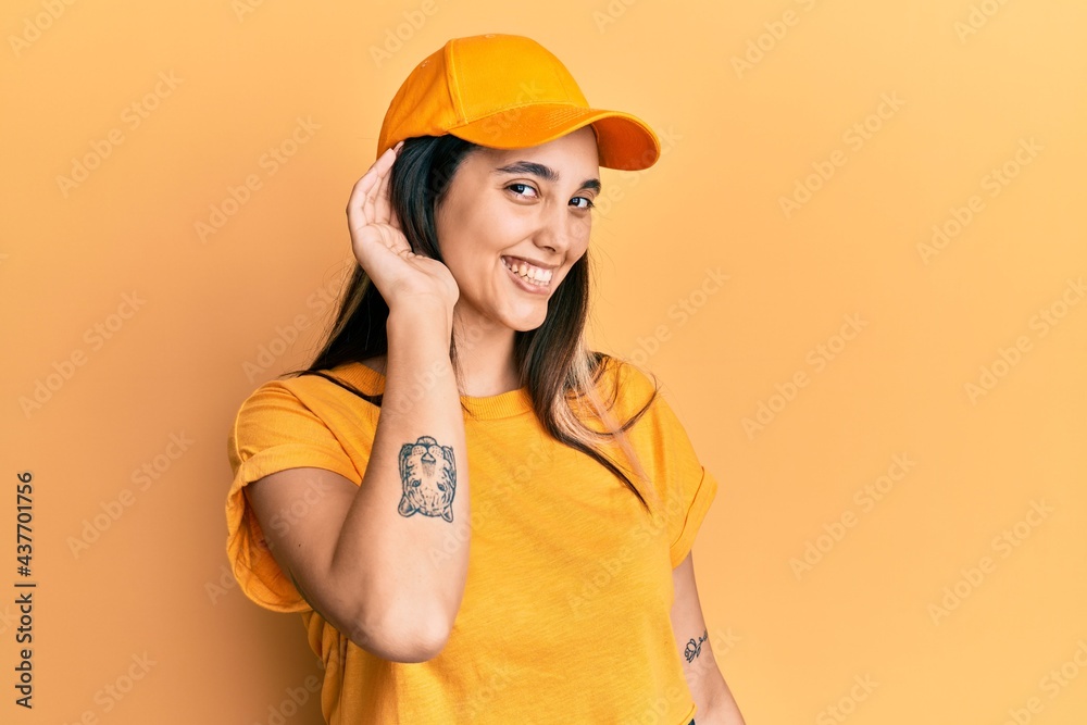 Young hispanic woman wearing delivery uniform and cap smiling with hand over ear listening an hearing to rumor or gossip. deafness concept. Young hispanic woman wearing delivery uniform and cap smiling with hand over ear listening an hearing to rumor or gossip. deafness concept.
