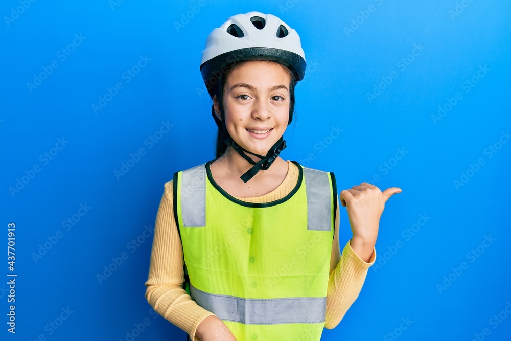 Beautiful brunette little girl wearing bike helmet and reflective vest smiling with happy face looking and pointing to the side with thumb up. Beautiful brunette little girl wearing bike helmet and reflective vest smiling with happy face looking and pointing to the side with thumb up.