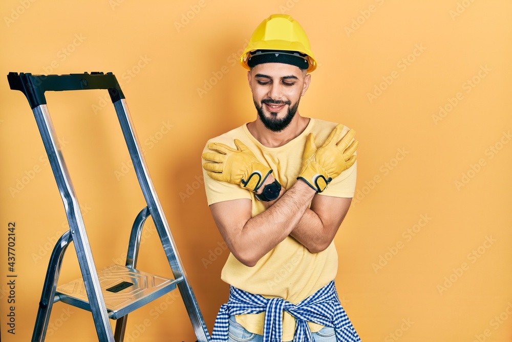 Handsome man with beard by construction stairs wearing hardhat hugging oneself happy and positive, smiling confident. self love and self care Handsome man with beard by construction stairs wearing hardhat hugging oneself happy and positive, smiling confident. self love and self care